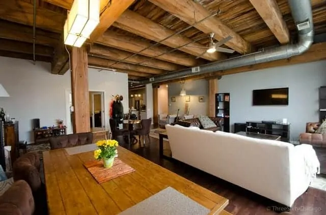 Photo of a loft condo with massive timber beams, exposed ductwork, a white couch and long wooden dining table with interesting rectangular light fixture.