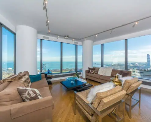 Interior of a luxury Chicago condo showing an open concept living room and kitchen with floor-to-ceiling windows overlooking the downtown city skyline.